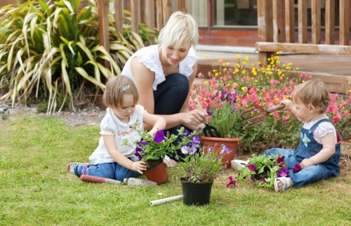 A caring woman with short blonde hair, dressed in a white top, is seated on a well-maintained lawn in a residential garden in Wallington, engaged in planting and tending to a variety of flowering plants in small pots and flower beds. The garden features a lush, green grass area with evenly cut turf, bordered by ornamental shrubs with long, yellow-green leaves in the background, alongside a wooden deck or patio that extends to the house. The flower beds contain vibrant pink, purple, and yellow blossoms, adding colour to the natural scene, with some plants in individual containers placed on the grass. The environment appears bright and sunny, suggesting fair weather, with natural daylight illuminating the scene and casting soft shadows. This outdoor space demonstrates typical features of a well-kept domestic garden suitable for family activities and outdoor gardening maintenance, aligning with services offered by Gardening Services Wallington in the SM postcode area.