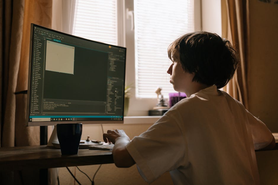A person with short dark hair sitting at a wooden desk indoors, working on a desktop computer with a large monitor displaying a programming or coding interface. The room is softly lit by natural light