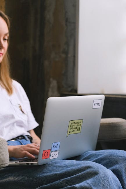 A person with long brown hair, wearing a white shirt, is sitting outdoors on a grey bench or chair, operating a silver laptop that is decorated with colorful stickers including a yellow binary code st