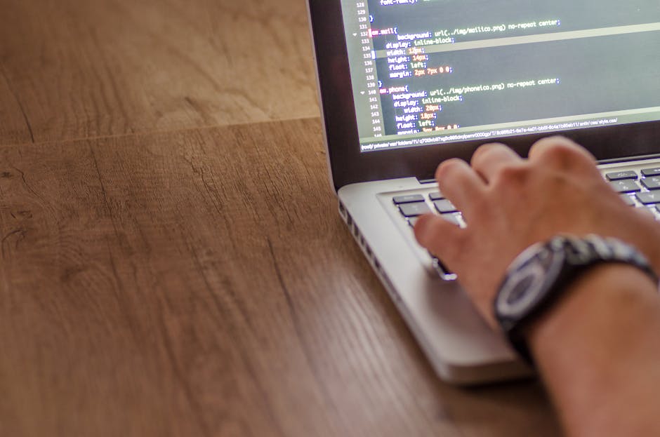 A person with long brown hair, wearing a white shirt, is sitting outdoors on a grey bench or chair, operating a silver laptop that is decorated with colorful stickers including a yellow binary code st